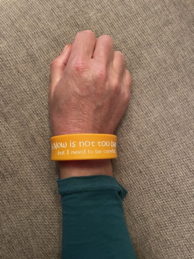 Hand and wrist shown wearing red and orange double sided silicone wristband reading 'now is not too bad but I need to be careful'