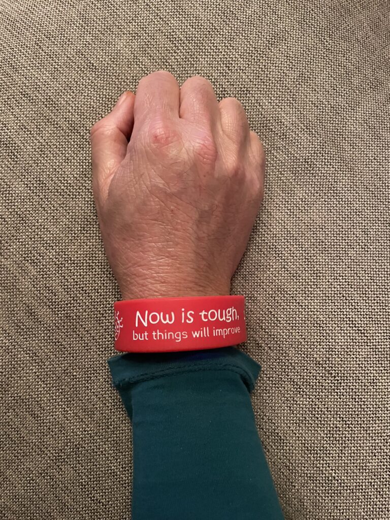 Hand and wrist shown wearing red and orange double sided silicone wristband reading 'now is tough but things will improve'