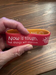 Hand holding red and orange double sided silicone wristband reading 'now is tough but things will improve'