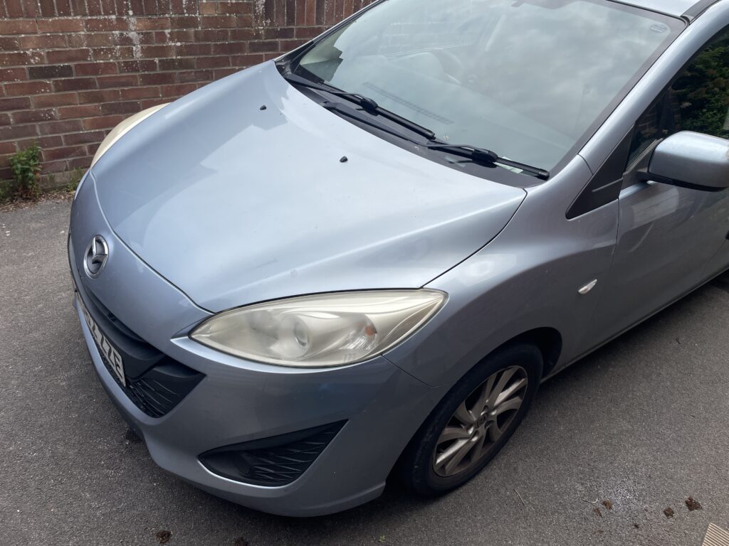 Front view of a light blue Mazda parked on a driveway next to a brick wall