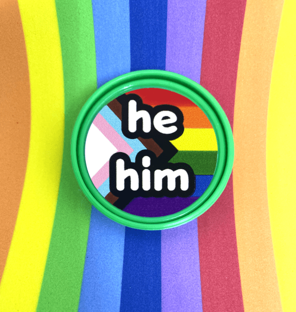 Pronoun pop badge with Pride flag backgrounds – he/him – from Sensory Oojamabobs on a rainbow striped background.