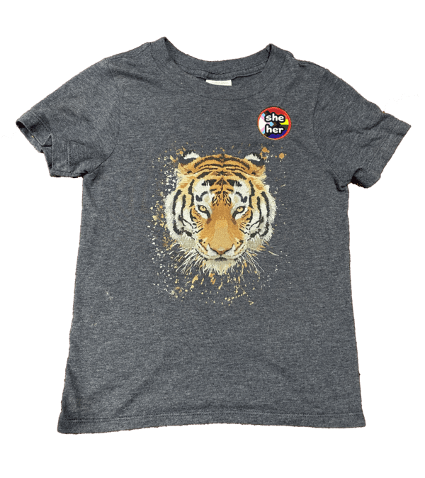 Pronoun pop badge with Pride flag backgrounds – she/her – from Sensory Oojamabobs on a navy child's t-shirt with a tiger on it