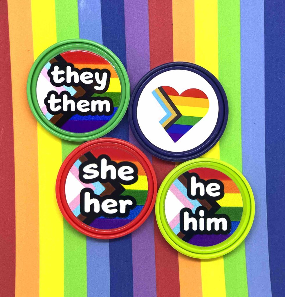 Pronoun pop badges with Pride flag backgrounds – they/them, she/her, he/him – from Sensory Oojamabobs on a rainbow striped background.